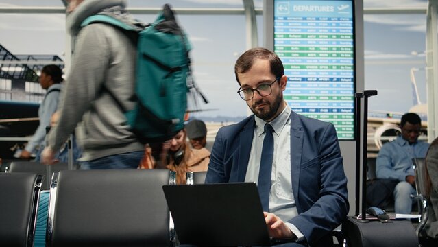 Freelancing man works on a laptop while waiting for an airport departure gate to open, completing remote tasks during international travel and preparing for his next flight from the airport lounge.