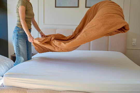 Woman making bed and spreading brown duvet over mattress with fitted sheet in bedroom