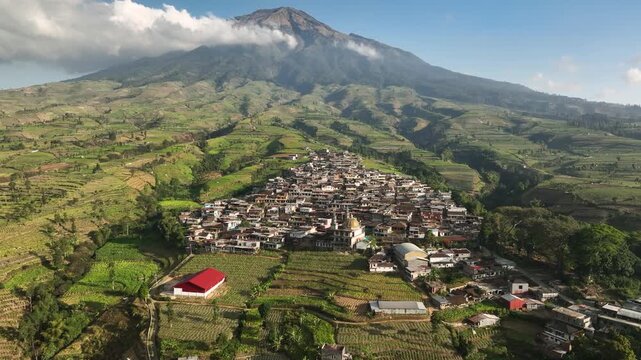 Aerial view of Kruwisan Village nestled among lush terraced agricultural fields at the base of a towering volcano in Kledung, Central Java, Indonesia.