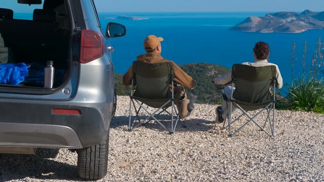 Couple enjoying scenic ocean view during road trip. Rear view of a man and a woman sitting on camping chairs next to their suv, peacefully enjoying a beautiful panoramic seascape with islands
