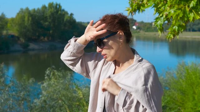 Woman suffering from heat stroke near lake during summer. Woman experiencing heat stroke symptoms near lake, sweating profusely, removing clothing while struggling with intense summer temperature