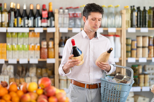 Man in light shirt took some varieties of wine from shelf. Client compare heady and new red wine items package. Customer examines contents of package, choose wine