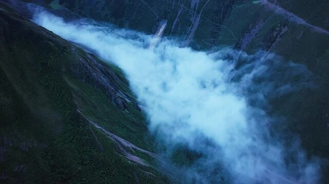 High-angle view captures thick white clouds drifting through a steep mountain valley under dim twilight illumination.