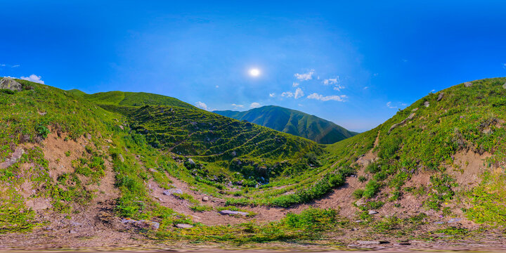 360-degree equirectangular panorama captures green mountainous landscape at surface level. Features include rolling hills, terraced slopes, rocky ground, and distant mountain ranges under a clear blue
