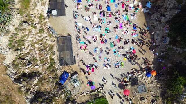 Aerial view of Polignano a Mare beach with people sunbathing on the pebbly shore of Lama Monachile beach surrounded by rocky cliffs in Polignano a Mare, Apulia, Italy.