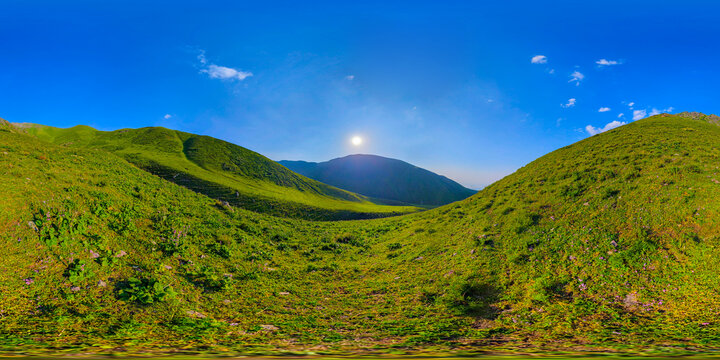 Full spherical panorama captures a mountainous landscape in Kyrgyzstan. Rolling green hills with purple flowers lead to distant mountains under a clear blue sky, illuminated by bright sunlight.