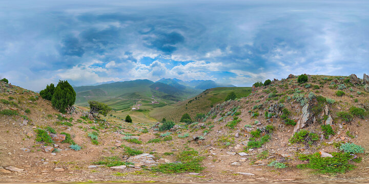 Full spherical panorama captured at surface level in Kyrgyzstan's mountainous region. Scene features rocky terrain, rolling green hills, and distant layered mountains under a cloudy sky.