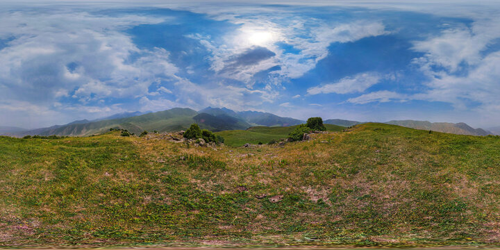 Full spherical panorama captured at ground level in Kyrgyzstan, showcasing rolling hills and distant mountains under a blue sky with clouds. Terrain features a mix of green and dry grass, with