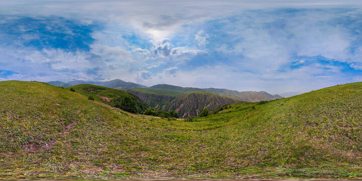 360-degree panoramic view captures rolling green hills and rugged mountains in Kyrgyzstan. Sparse vegetation and rocky outcrops enhance the natural beauty under a partly cloudy sky.