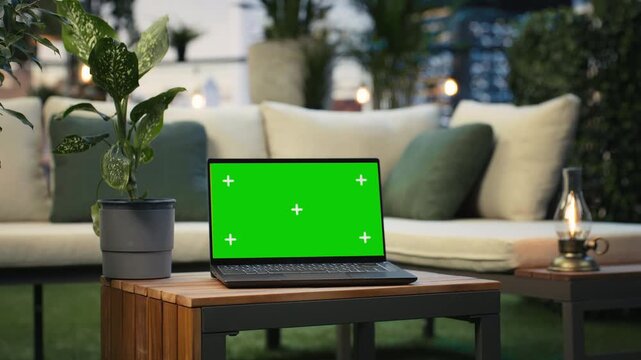 Mockup laptop with isolated screen on a rooftop lounge table beside lush greenery, showing a close-up chroma key notebook display in a terrace workspace with city skyline in the background.