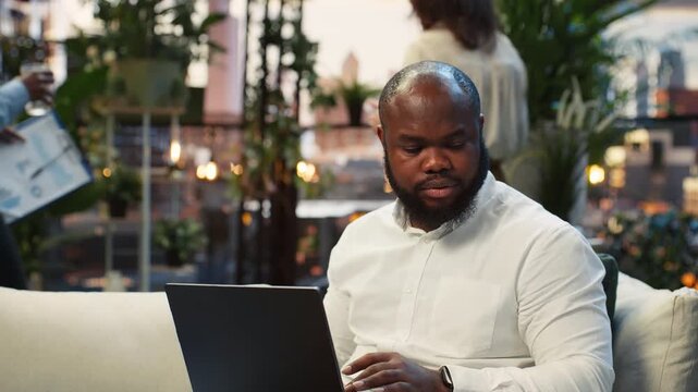Happy businessman reads laptop information while working on a rooftop terrace, researching and analyzing project data during a focused work session.