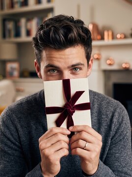 Smiling man holding a beautifully wrapped Christmas gift white envelope with a festive ribbon in a cozy living room