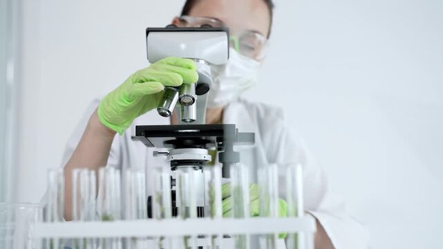 Scientist in a lab coat and green gloves carefully adjusting a microscope to focus on plant samples in test tubes, performing detailed research in a sterile laboratory environment