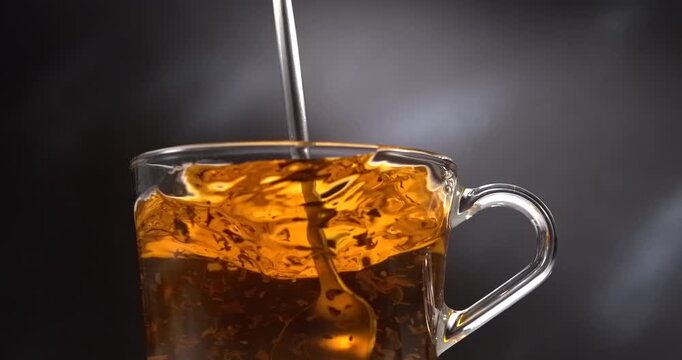 Hot black tea stirring with spoon in glass cup with steam on black background in slow motion