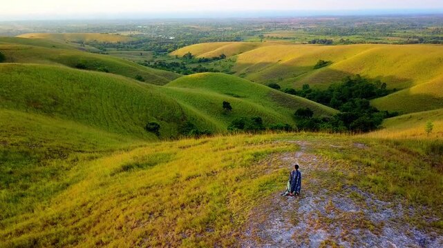 Aerial view of Bukit Wairinding with rolling green hills under golden hour light and a person standing on the grassy ridge in Nggaha Oriangu, East Nusa Tenggara, Indonesia.