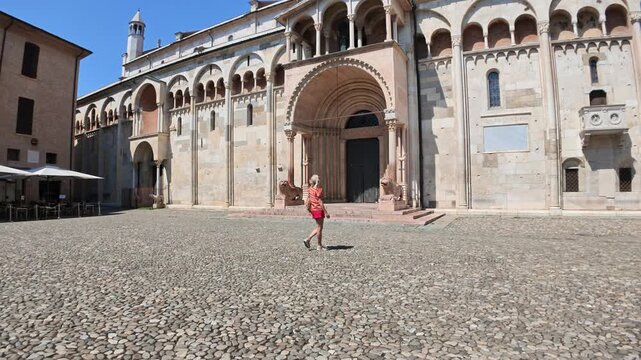 Woman in red walking on the historic cobblestone piazza, admiring the ancient Romanesque architecture of Modena Cathedral, a famous UNESCO world heritage site of Modena city in Italy