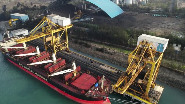 Aerial view of the cargo ship PLN-BARTERA ADHIGUNA being loaded with coal by large yellow industrial cranes at a port in Adipala, Central Java, Indonesia.