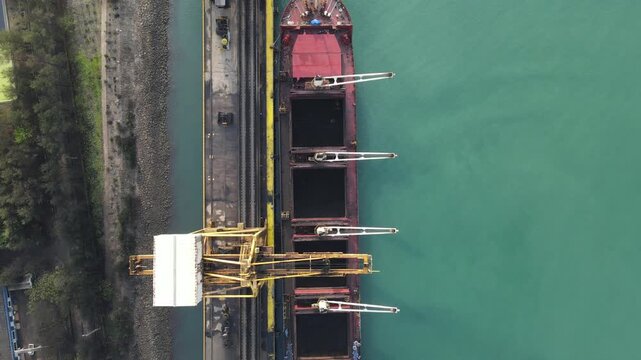 Aerial view of a large cargo ship docked at a port with industrial cranes and turquoise water in Adipala, Central Java, Indonesia.
