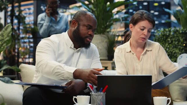 Businesspeople analyze laptop information together during rooftop workspace collaboration, comparing data on a notebook in an urban lounge above offices.