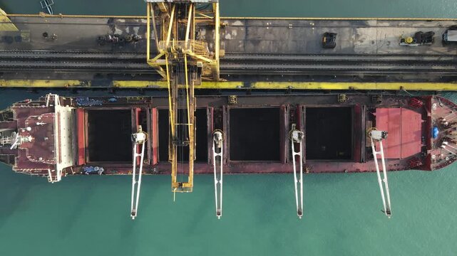 Aerial view of a large cargo ship being loaded with coal by a yellow crane at the port in Adipala, Central Java, Indonesia.