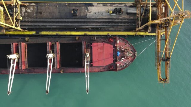 Aerial view of a large cargo ship docked at a port with a yellow crane and turquoise water in Adipala, Central Java, Indonesia.