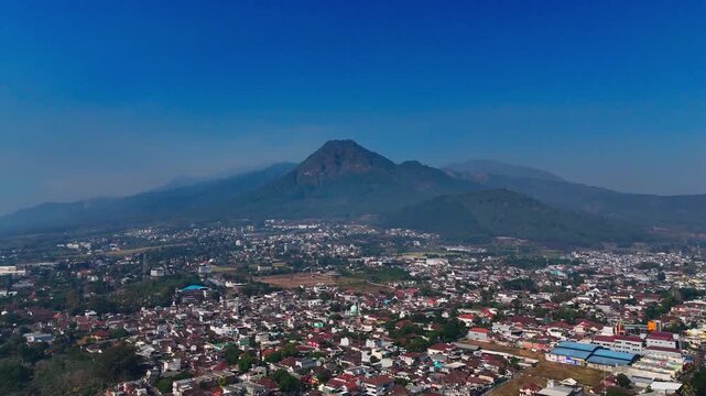 Aerial view of Mount Panderman towering over the urban landscape of Batu city with dense residential rooftops and lush surroundings in Batu, East Java, Indonesia.