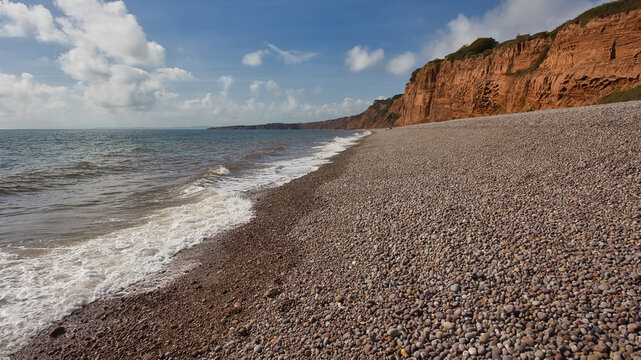 A wide landscape shot captures gentle waves breaking on a sprawling pebble beach, bordered by dramatic red sandstone cliffs beneath a cloudy blue sky.