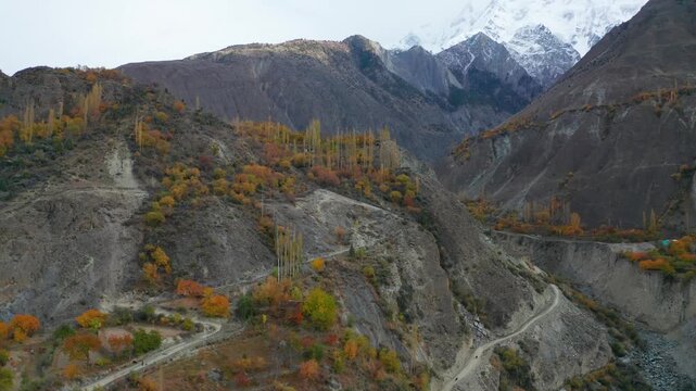 Aerial view of Sost Valley with winding mountain roads, rocky slopes, and vibrant autumn foliage under snow-capped peaks in Sost, Gilgit, Pakistan.