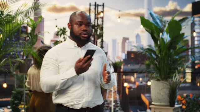 Businessman sends smartphone voice notes at a rooftop networking event while colleagues talk nearby, responding to business messages during the gathering.