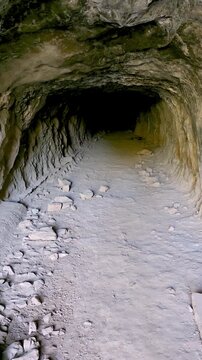 POV smooth camera moving forward into a dark cave entrance carved into the rock. View from outside daylight entering a deep underground stone passage with scattered rocks on the ground.