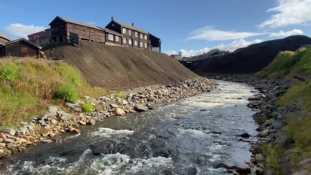 A river with stony banks, flowing next to an old wooden building, embedded in a hilly landscape, mining spoil heap, copper ore, Unesco World Heritage Site, R&ouml;ros, Norway