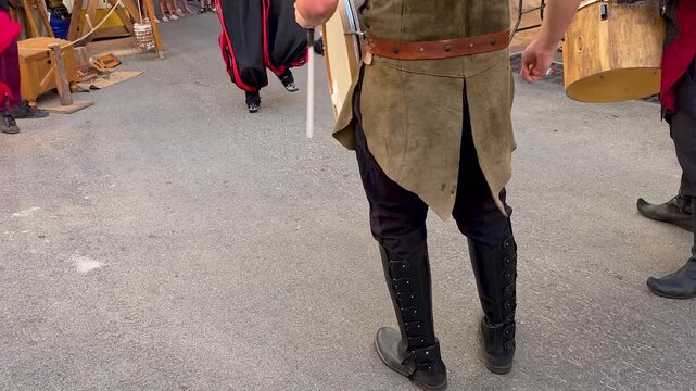 Medium shot of street musicians in traditional medieval clothing and leather boots playing wooden drums during a festive procession at a Spanish harvest festival.