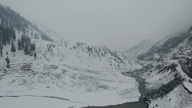 Aerial view of Naran Kaghan valley covered in snow with a winding river and road under a cloudy winter sky in KPK, Pakistan.