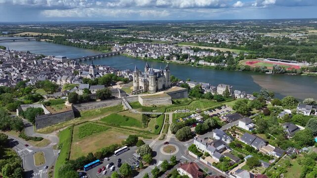 Aerial view of the historic Ch&acirc;teau de Saumur with its distinctive round corner towers and bright facades, surrounded by well-kept green spaces under a slightly cloudy summer sky, with the town of S