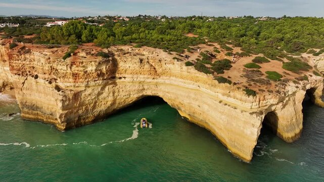 Drone view of Benagil Cave cliffs in Algarve, Portugal, with a tourist boat near limestone sea caves, turquoise Atlantic water, and rugged coast. Slide trucking shot.