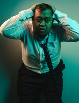 A terrified adult Asian male stares wide eyed with hands near his head, illustrating extreme shock and workplace panic. The chubby corporate employee wears an untidy white long sleeve