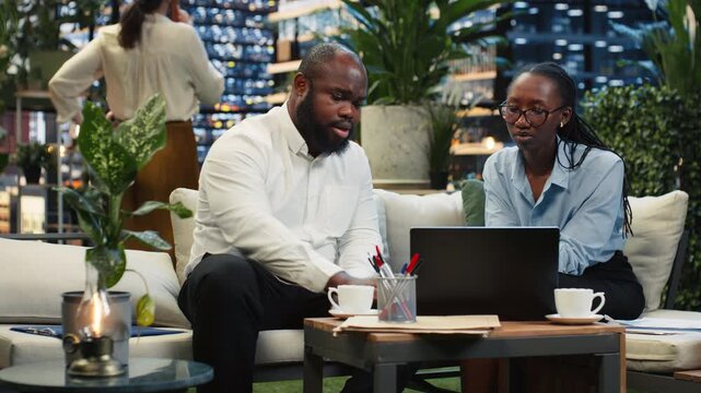 Company colleagues review laptop data together on a rooftop terrace, comparing notebook information and discussing research findings in the metropolitan lounge.