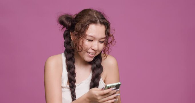 Young Kazakh woman smiling while using smartphone against pink background