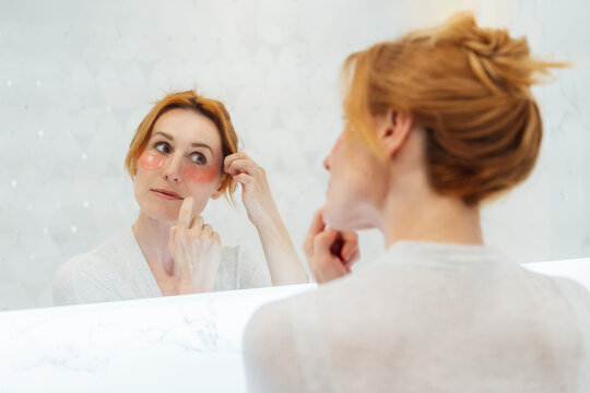 Self Cosmetology, skin care, face treatment, natural beauty concept. Middle aged woman in pajamas looking in mirror and applying pink moisturizing patches on eyes in the bathroom. Home Beauty routine.