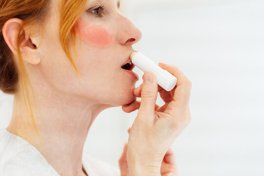 Close up side view Middle aged woman applies lip balm looking in mirror in white bathroom. SPF Lip Balm for lips kin protection. Home beauty treatment and skin care routine.