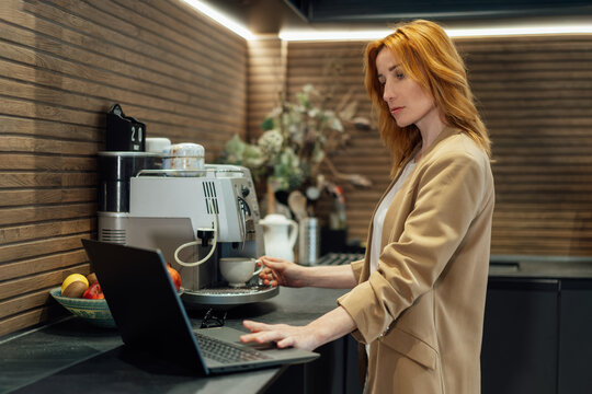 Middle aged businesswoman making morning coffee in office kitchen and checking working plans on laptop . Female preparing hot drink with espresso machine. Corporate lifestyle daily routine concept.