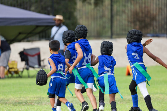 A group of young children wearing blue uniforms, soft protective helmets, and green flags run together during a youth flag football game on a grassy field.