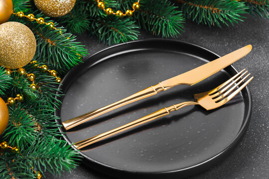 Elegant Christmas table setting with gold cutlery on a black plate, surrounded by evergreen branches, golden ornaments, beads and pinecones on a dark textured background
