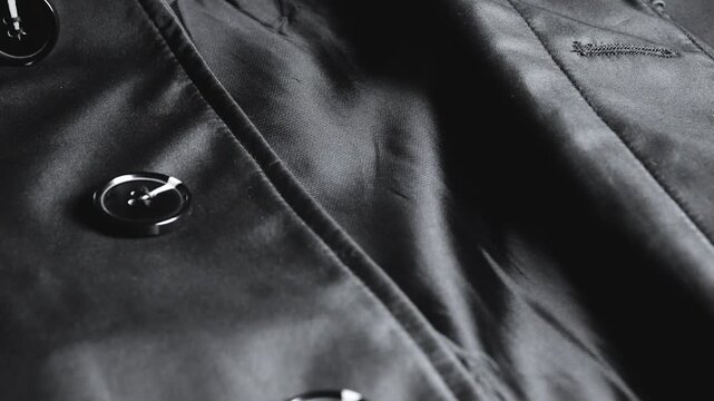 A close-up of a black satin trench coat with large round buttons showcases smooth waterproof fabric, neat seams, and elegant structural details of classic outerwear.
