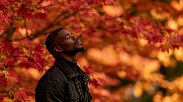 a man standing in a forest during autumn. He is captured in a side profile, looking upwards with his eyes closed, seemingly lost in thought. The man wears a dark jacket and a black turtleneck sweater.