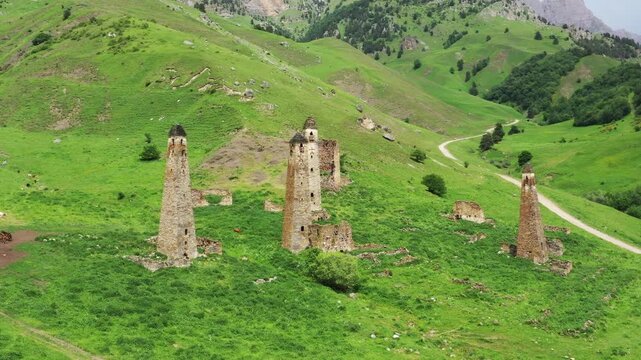 Aerial view of the ancient Caucasus Towers surrounded by lush green hills and a winding dirt road in Ingushetia, Russia.