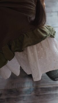 Young girl puts on black rain boots indoors. focus is close up of her lower body and hands as girl pulls up skirt revealing white dress with olive green top layer underneath. wooden floor