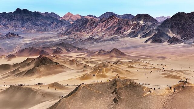 High-angle panorama captures tourists exploring vast sand dunes and rugged mountain ridges under a clear blue sky in Qinghai.