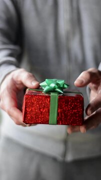 Close-up of hands offering red gift box with green ribbon toward the camera, person giving gift to friend or relative