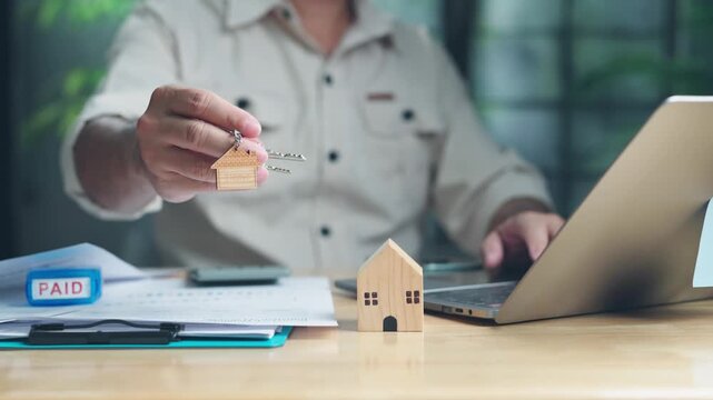 Midsection man holding house keychain beside wooden house model, paid stamp, laptop on desk while finalizing home mortgage payment indoors.